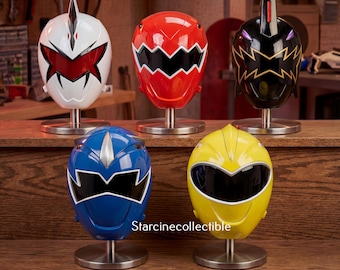 Peut inclure: Cinq casques Power Rangers colorés, blancs, rouges, noirs, bleus et jaunes, chacun avec des visières uniques, montés sur des socles argentés. Le texte "Starcinecollectible" est visible en bas à droite.