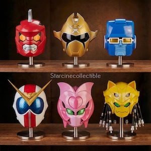 May include: Six character helmets in various colours, including red, gold, blue, white, pink, and yellow, displayed on metal stands. The helmets feature diverse designs, such as angular shapes and visors. The text "Starcinecollectible" is visible.