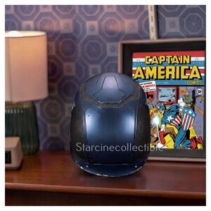 May include: A dark blue Captain America helmet with black accents, displayed on a wooden surface. The helmet is next to a framed Captain America comic book cover. The background features a patterned wall and a lamp.