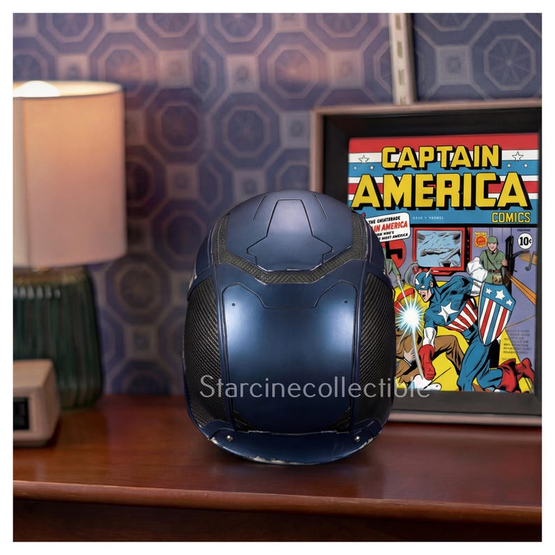 May include: A dark blue Captain America helmet with black accents, displayed on a wooden surface. The helmet is next to a framed Captain America comic book cover. The background features a patterned wall and a lamp.