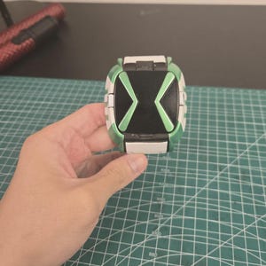 May include: A green, black, and white toy watch with an X-shaped design on the face. The watch is held in a hand and appears to be made of plastic. The watch has a rectangular shape with rounded edges.