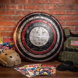 May include: A large, circular shield with a distressed metal finish, featuring concentric rings of black, silver, and red. Comic books with the text "USAGENT" and a helmet are visible. A tactical vest and gloves are also present.