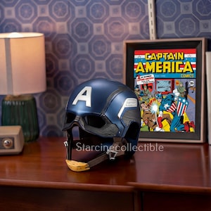 May include: A navy blue Captain America helmet with a white "A" on the front. The helmet has a black visor and a yellow chin strap. A framed "Captain America Comics" print is in the background.