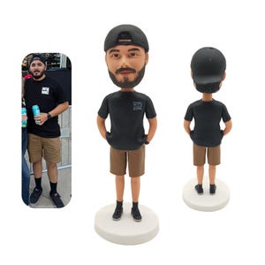 Op de afbeelding: Een gepersonaliseerd bobblehead-beeldje van een persoon die een zwart T-shirt draagt met een wit logo, een bruine korte broek en een zwarte baseballpet. Het beeldje staat op een witte basis.