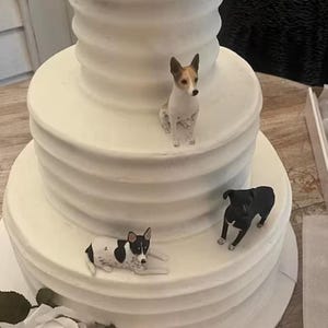 Può includere: Una torta nuziale bianca a tre piani decorata con tre piccole statuine di cani. Il cane in alto è marrone e bianco, il cane al centro è nero e il cane in basso è nero e bianco.
