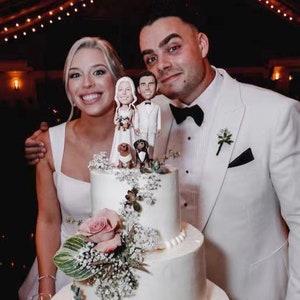 May include: A white wedding cake with a white fondant topper featuring a bride and groom, two dogs, and a floral arrangement. The cake is decorated with white flowers and greenery. The couple is smiling and holding hands. The cake is on a table with a white tablecloth.