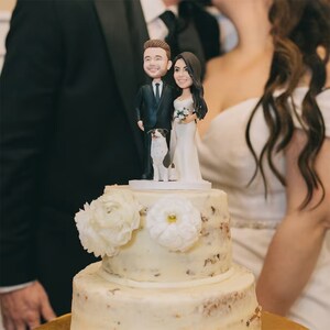 May include: A two-tiered wedding cake with white frosting and a topper featuring a bride and groom figurine with a dog. The cake is decorated with white flowers.