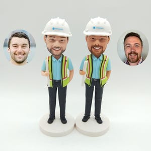 Peut inclure: Deux figurines de tête de chantier personnalisées portant des chemises bleues, des gilets verts et des casques blancs avec le logo "OpenSpace". Chaque figurine tient un plan roulé.