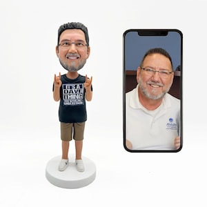 Op de afbeelding: Een aangepaste bobblehead van een man met een bril, een zwart t-shirt met de tekst "IT'S A DAVE THING YOU WOULDN'T UNDERSTAND", een kaki korte broek en witte sneakers. Een smartphone toont een foto van dezelfde man.