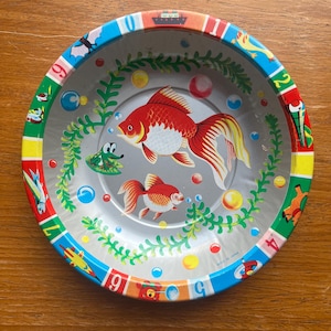 以下が含まれることがあります： カラフルなデザインのヴィンテージブリキのおもちゃのボウル。ボウルは、金魚、泡、水生植物のイラストが描かれたシルバーのインテリアが特徴です。リムは数字とさまざまな漫画の画像で装飾されています。ボウルは日本で作られています。