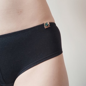 May include: Solid black underwear featuring a small, rectangular tan-colored fabric tag on the side waistband. The tag displays a dark green graphic of a leaf. The underwear has a simple, clean design with visible stitching along the top edge.
