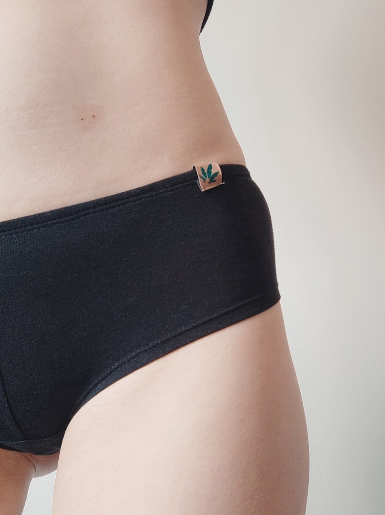 May include: Solid black underwear featuring a small, rectangular tan-colored fabric tag on the side waistband. The tag displays a dark green graphic of a leaf. The underwear has a simple, clean design with visible stitching along the top edge.