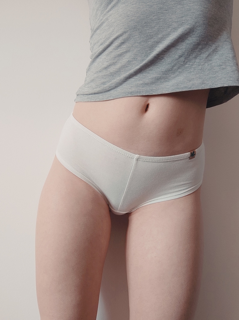 May include: A pair of solid white cotton-style briefs featuring a small, rectangular brown fabric tag on the right hip. The underwear has a clean, simple design with a standard waistband and leg openings, paired with a grey short-sleeved shirt.