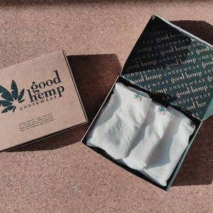 May include: A brown cardboard box with the text good hemp UNDERWEAR and a leaf graphic, next to an open box containing three pairs of white underwear. The inside lid of the open box features a repeating pattern of the text good hemp UNDERWEAR.