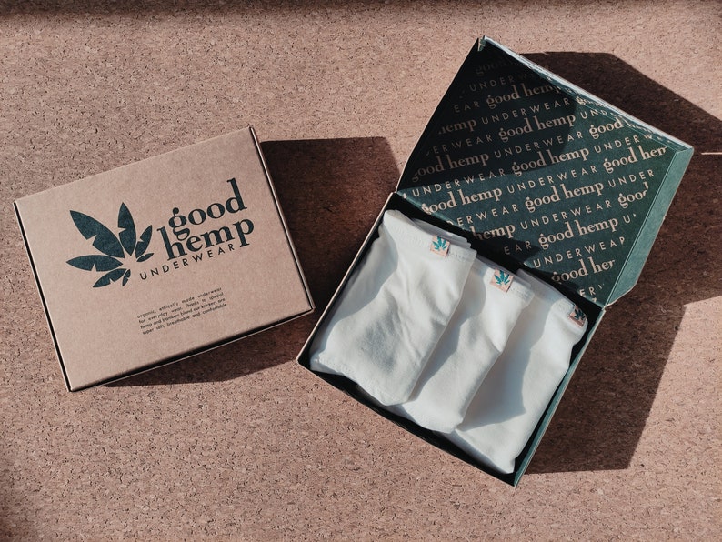 May include: A brown cardboard box with the text good hemp UNDERWEAR and a leaf graphic, next to an open box containing three pairs of white underwear. The inside lid of the open box features a repeating pattern of the text good hemp UNDERWEAR.