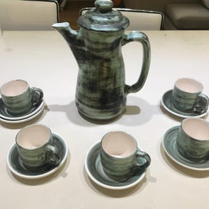 Puede incluir: Una cafetera de cerámica vintage con un esmalte verde y negro, junto con seis tazas y platillos a juego. Las tazas y los platillos tienen un esmalte verde y negro similar.