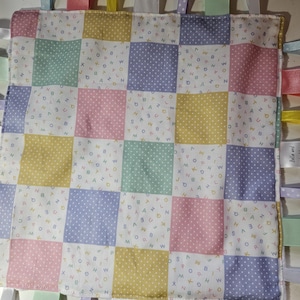 May include: A pastel-coloured baby tag blanket with a patchwork design. Each square is a different colour, with polka dots or letters. Ribbons in various colours are attached around the edges. The tag says "Bella and".