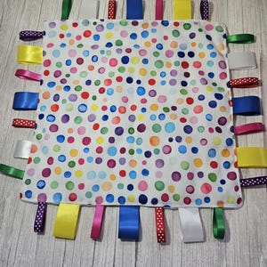 May include: A square fabric sensory blanket featuring a white background with a multicoloured polka dot pattern. The edges are adorned with numerous loops of satin and grosgrain ribbons in various colours including blue, yellow, pink, green, and white.
