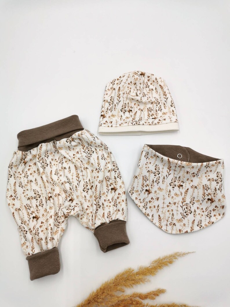May include: A baby outfit set with a brown and white floral print. The set includes a pair of pants, a hat, and a bib.