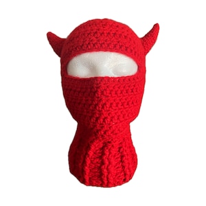 Puede incluir: Balaclava de punto rojo con cuernos. La balaclava tiene un agujero para los ojos y un cuello largo.
