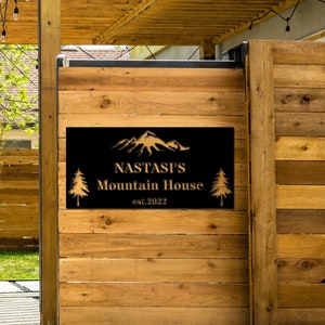 Könnte beinhalten: Ein schwarzes Metallschild mit goldenen Buchstaben, auf dem "Nastasi's Mountain House est. 2022" mit einer Bergkette und Kiefern steht. Das Schild ist an einem Holzzaun befestigt.