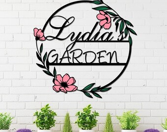 May include: Black metal garden sign with the name "Lydia's" and the word "GARDEN" in a circular design. Pink flowers and green leaves decorate the sign. The sign is mounted on a white brick wall above a wooden shelf with potted plants.