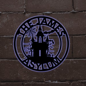 May include: A black metal sign with a spooky haunted house design and the text "The James Asylum".