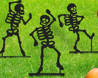 May include: Black metal skeleton yard decorations in dancing poses, set in green grass with orange pumpkins. The skeletons have detailed rib cages, skulls, and limbs, perfect for Halloween. The pumpkins add a festive touch.