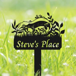 May include: Black metal garden stake featuring a detailed gecko design amidst foliage. The stake has the text "Steve's Place" cut out at the bottom. The stake is set in a grassy area with small white flowers.