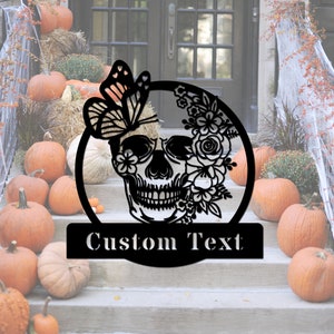 Może przedstawiać: Czarna metalowa dekoracja drzwi na Halloween z czaszką ozdobioną kwiatami i motylem. Czaszka ma kwiatowy wz&oacute;r i motyla z boku. Tekst "Custom Text" znajduje się pod czaszką.