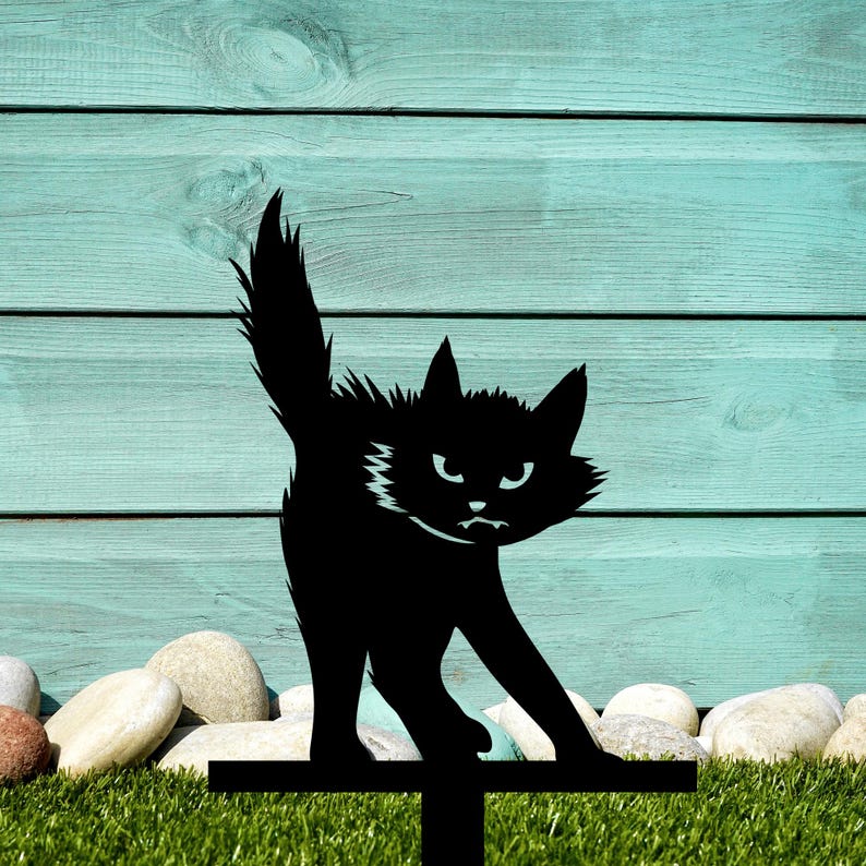May include: Black metal silhouette of a cat with an angry expression, standing on a metal stake. The cat is facing the viewer with its tail raised. The stake is placed in front of a green grassy area.