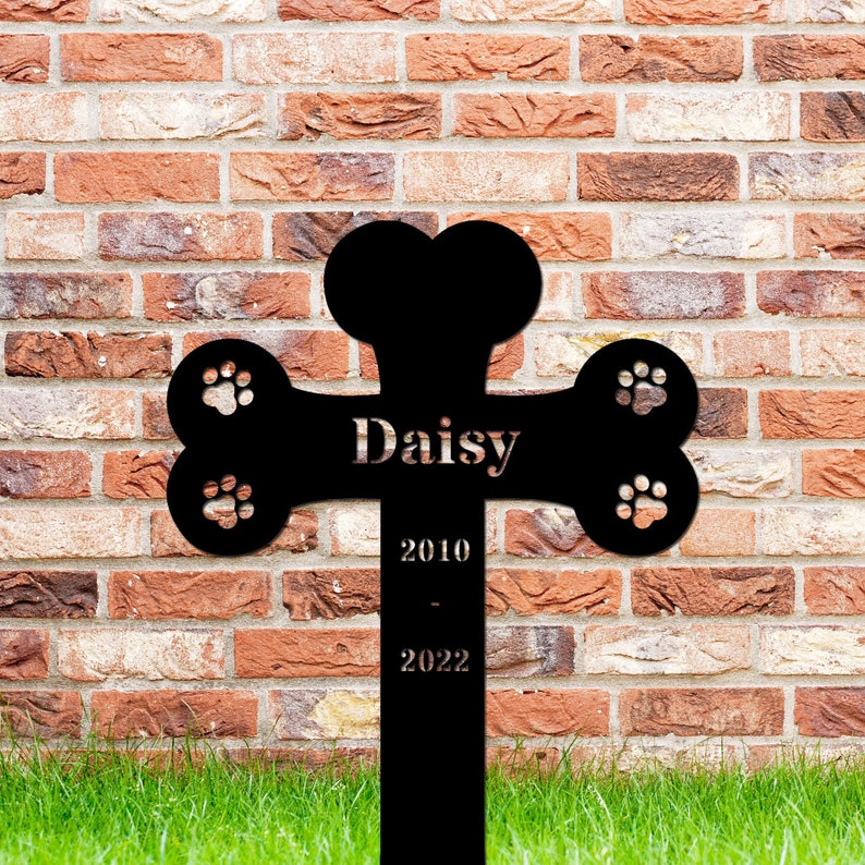 May include: Black metal pet memorial cross shaped like a bone with paw prints. The cross has the name "Daisy" and the dates "2010 - 2022" engraved on it.
