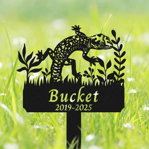 Może przedstawiać: Czarny metalowy palik ogrodowy z wzorem gekona w kropki. Palik zawiera imię "Bucket" i lata "2019-2025" w kolorze zielonym, na tle zielonej trawy i liści.