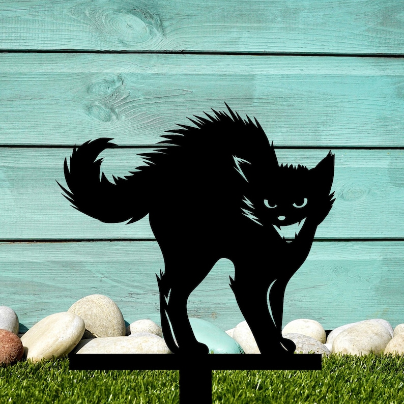 May include: Black metal silhouette of a cat with a menacing expression, standing on a metal stake. The cat is facing forward with its mouth open, showing sharp teeth. The stake is placed in a bed of green grass.
