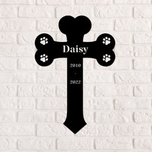 May include: Black metal pet memorial cross with a bone shape and paw prints. The cross has the name "Daisy" and the dates "2010 - 2022" engraved on it.