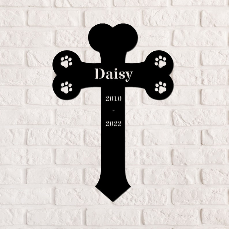 May include: Black metal pet memorial cross with a bone shape and paw prints. The cross has the name "Daisy" and the dates "2010 - 2022" engraved on it.