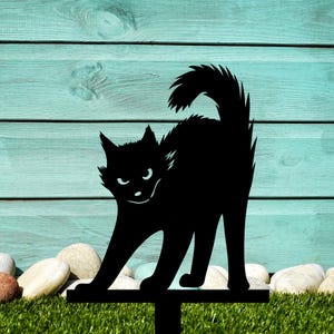 May include: Black metal silhouette of a cat with a menacing expression, standing on a metal stake. The cat is positioned against a light blue wooden background.