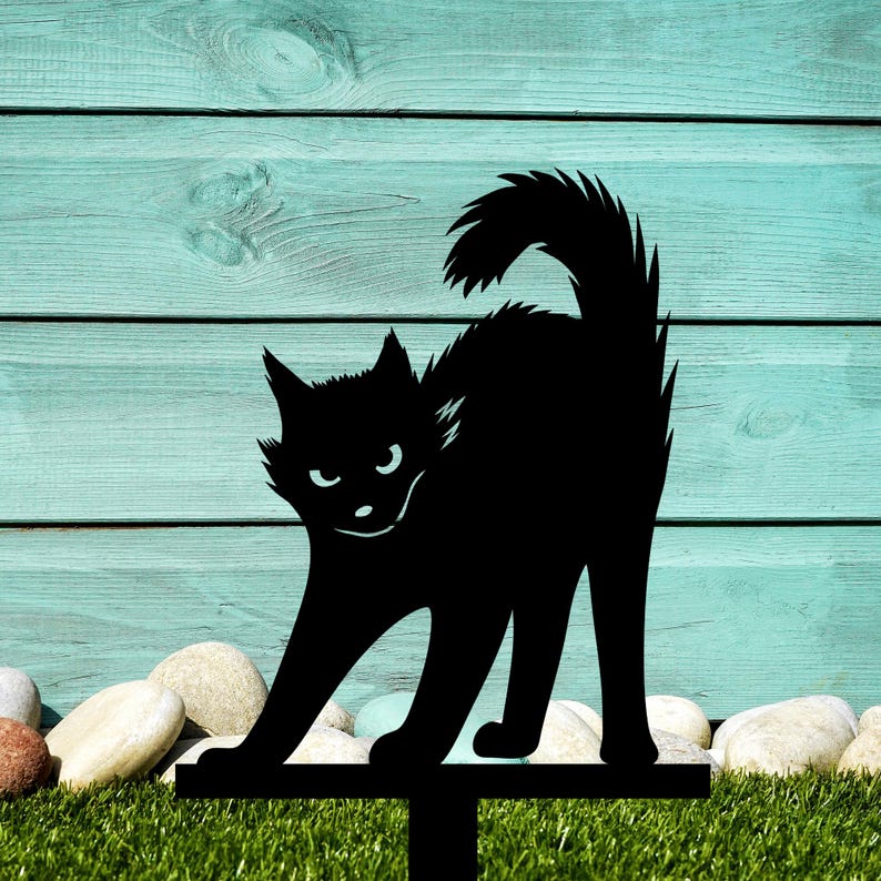 May include: Black metal silhouette of a cat with a menacing expression, standing on a metal stake. The cat is positioned against a light blue wooden background.