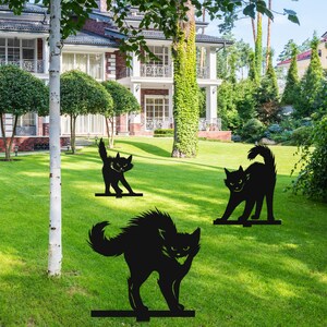 May include: Three black metal silhouette yard decorations of cats in various poses. The cats are standing on a green lawn in front of a house.