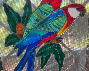 Könnte beinhalten: Zwei bunte Glas-Papageien, die auf einem Ast mit grünen Blättern sitzen. Die Papageien sind rot, blau, gelb und grün. Die Blätter sind grün.