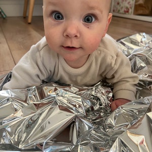May include: A baby wearing a beige onesie is lying on a silver crinkle blanket. The baby is looking directly at the camera with a curious expression.