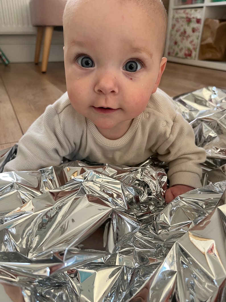May include: A baby wearing a beige onesie is lying on a silver crinkle blanket. The baby is looking directly at the camera with a curious expression.