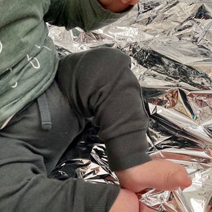 May include: A baby wearing a green long-sleeved shirt with white text and grey sweatpants sits on a silver foil blanket.