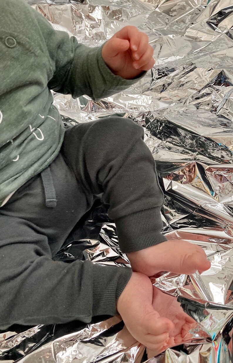 May include: A baby wearing a green long-sleeved shirt with white text and grey sweatpants sits on a silver foil blanket.