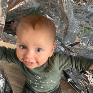 May include: A baby with blue eyes wearing a green long-sleeved shirt with a white plant graphic and the text "Little Ku..." on it. The baby is looking at the camera and smiling.
