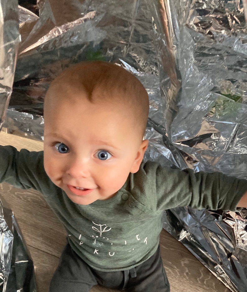 May include: A baby with blue eyes wearing a green long-sleeved shirt with a white plant graphic and the text "Little Ku..." on it. The baby is looking at the camera and smiling.