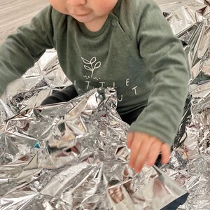 May include: A baby wearing a green long-sleeved shirt with a white plant graphic and the text "Little Out" is playing with a silver emergency blanket.
