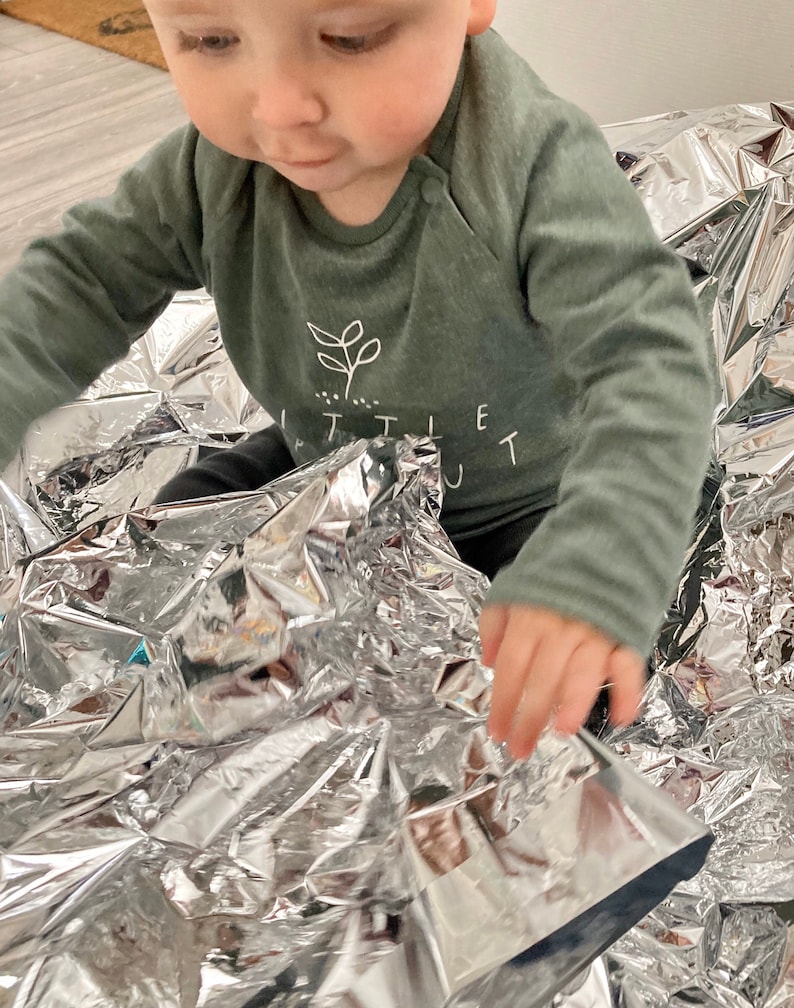 May include: A baby wearing a green long-sleeved shirt with a white plant graphic and the text "Little Out" is playing with a silver emergency blanket.