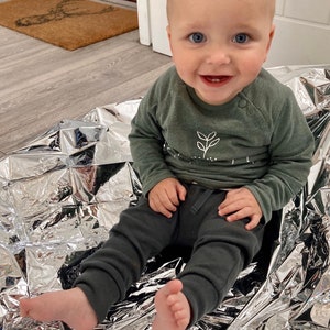 May include: A baby boy wearing a green long-sleeved shirt with a white plant design and dark grey trousers sits on a silver blanket. The baby is smiling and looking at the camera.