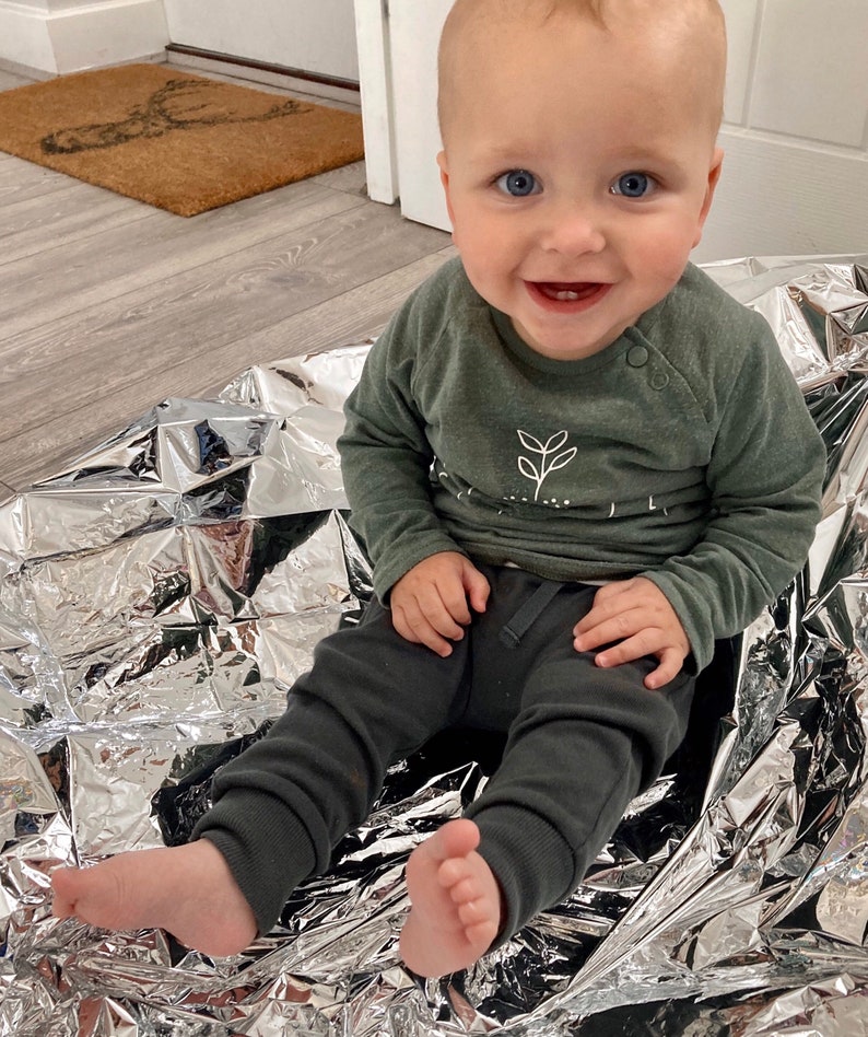 May include: A baby boy wearing a green long-sleeved shirt with a white plant design and dark grey trousers sits on a silver blanket. The baby is smiling and looking at the camera.